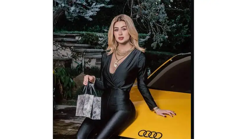 A blond influencer poses with a shopping bag and a yellow sportscar