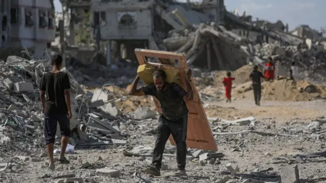 A man in the centre of the frame carries a broken yellow door on his back, other people walking around the rubble and ruins of bombed out homes in the background