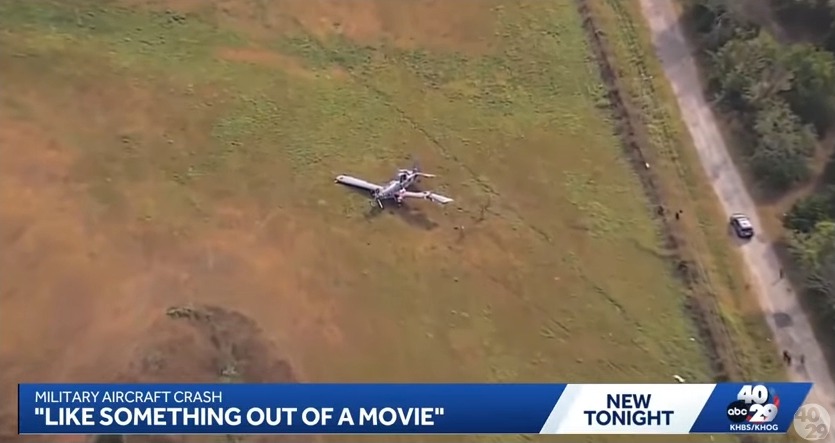 Aerial view of a military aircraft crashed in a field, with a news chyron reporting "MILITARY AIRCRAFT CRASH".