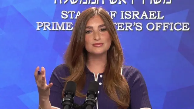 A woman speaking into two microphones on stands and gesturing with one hand up. The background behind her is blue